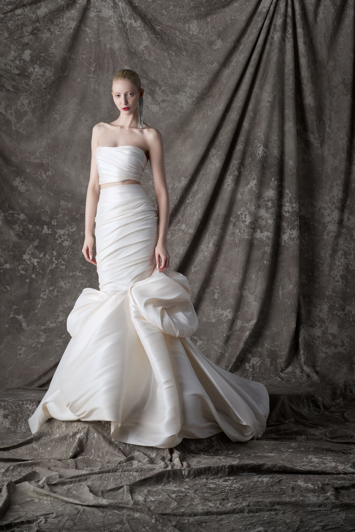 White glass fabric wedding dress with a large bow at trumpet hem on a textured gray backdrop